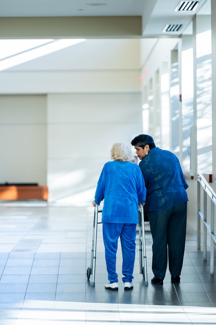 gallery-2 A nurse assists an elderly woman with a walker in a bright hospital corridor.