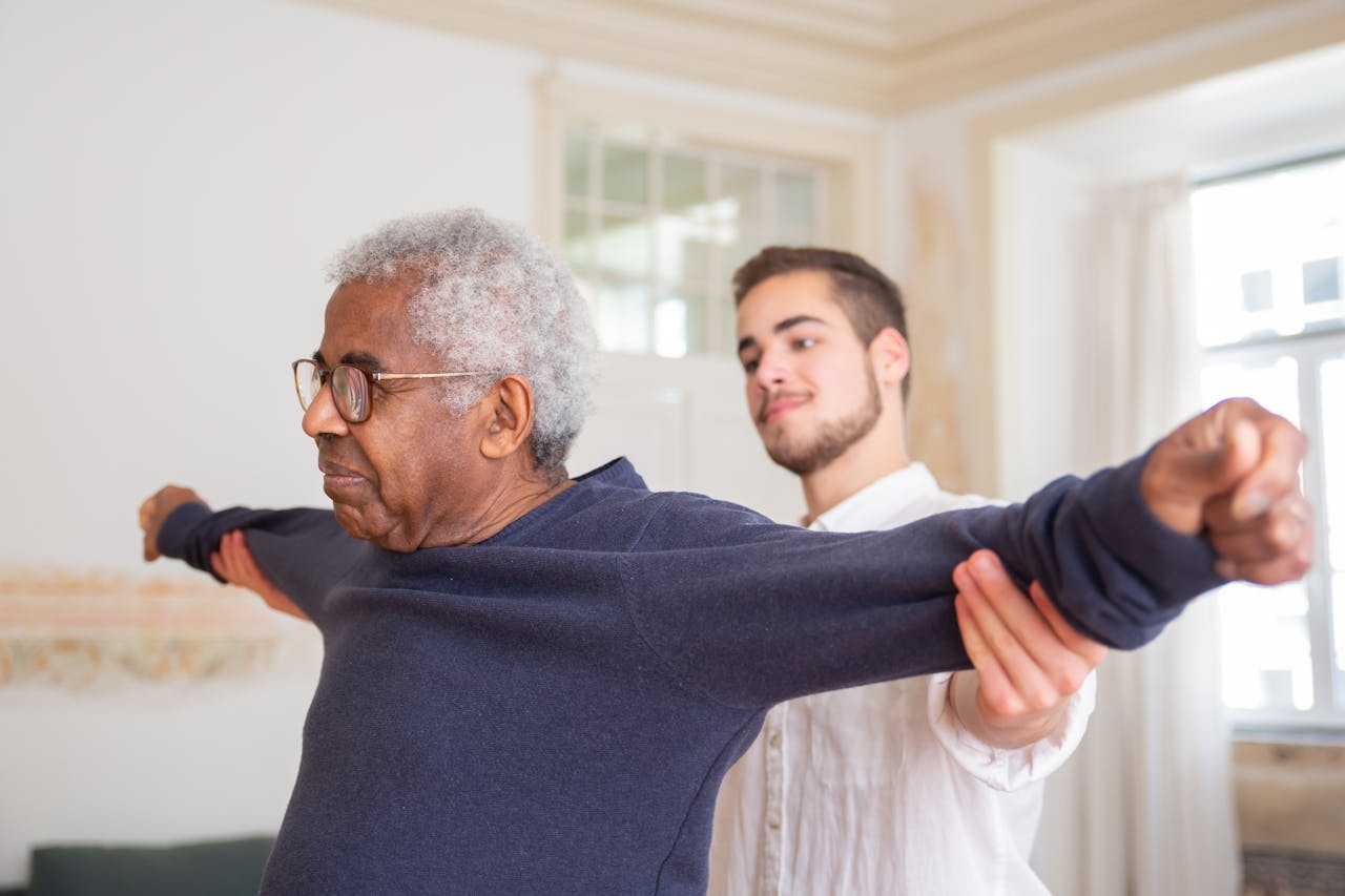 our-services-3 A senior man is assisted by a caregiver for stretching exercises indoors.